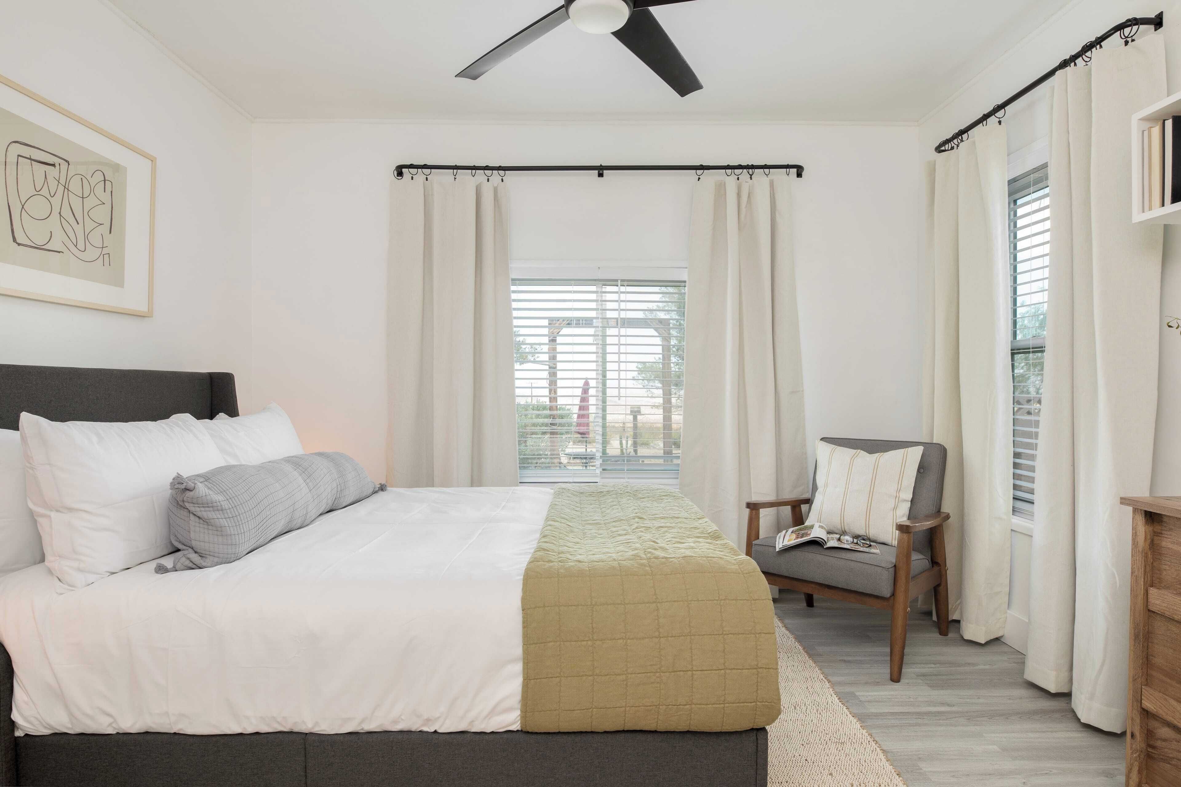 Une chambre à coucher avec un lit, un ventilateur de plafond, une fenêtre avec des rideaux, une chaise et une bibliothèque.