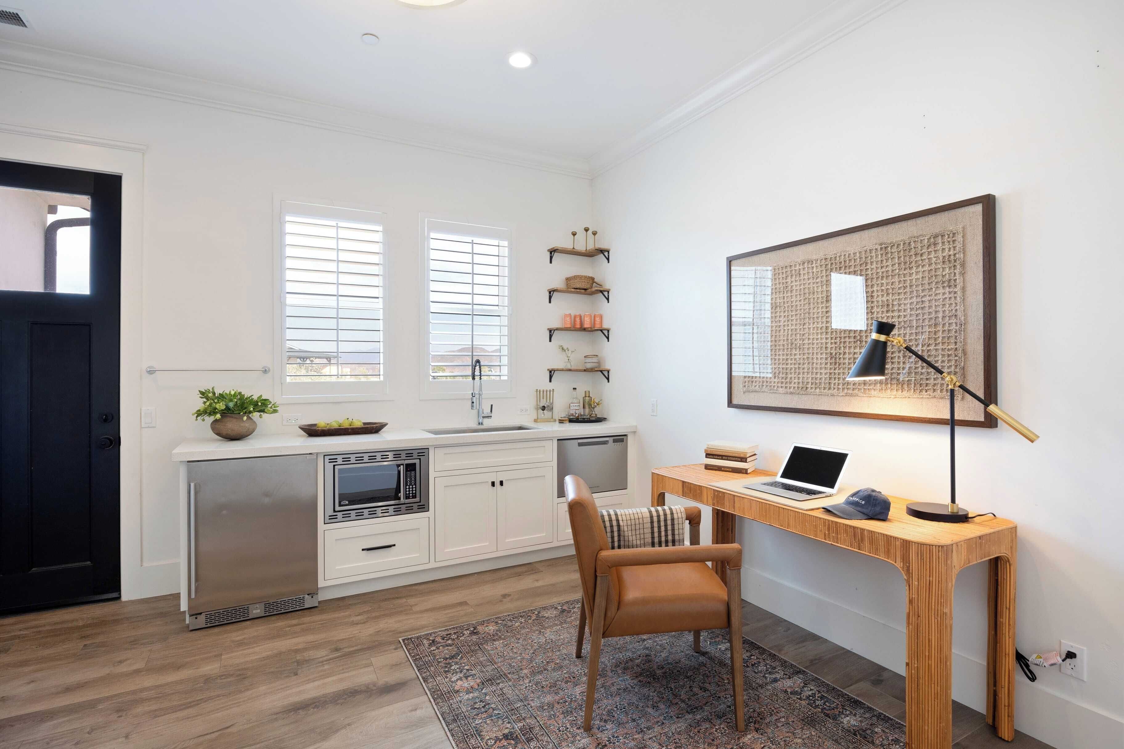 A modern kitchen with white cabinetry, a built-in oven, and a microwave. There is a wooden desk with a laptop and a lamp.