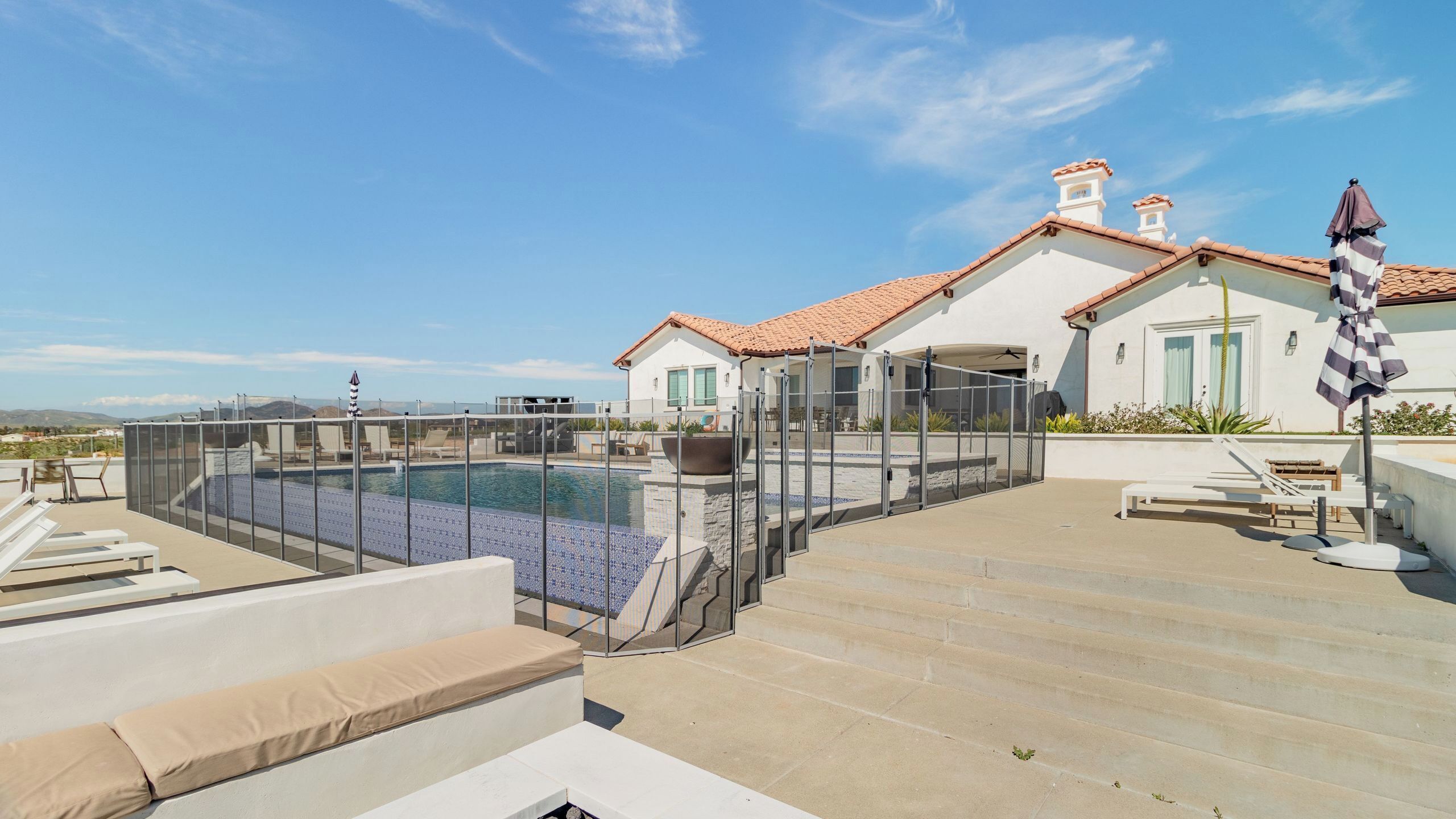 A rooftop pool with a glass enclosure, outdoor seating, and a view of a white building with a tiled roof.
