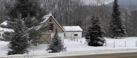 A snow-covered house with a red door and windows, surrounded by snow-laden trees and a fence.