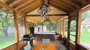 Una terraza cubierta de madera con una mesa central y bancos, un jacuzzi y vistas a una casa y árboles.