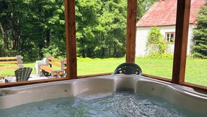 Un jacuzzi bajo una pérgola de madera con vistas a un paisaje verde y un edificio blanco con tejado rojo.