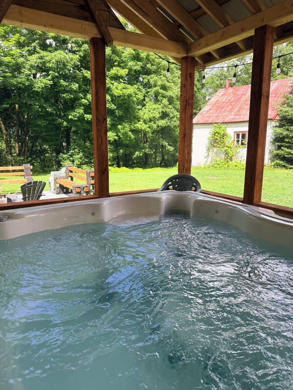 A hot tub under a wooden pergola with a view of a green landscape and a white building with a red roof.