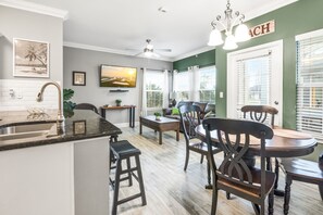 A modern kitchen with a central island, wooden dining table, and chairs. There is a ceiling fan, a TV mounted on the wall, and a window with blinds.