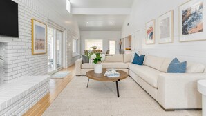 A modern living room with a white brick fireplace, a beige sofa, a round wooden coffee table, and framed artwork on the walls. - Twin Fin by Avantstay Walk to the Beach - Chic 30A Retreat (Panama City Beach)