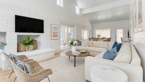 A spacious living room with a white brick wall, a flat-screen TV, wicker furniture, a beige sofa, and a ceiling fan. - Twin Fin by Avantstay Walk to the Beach - Chic 30A Retreat (Panama City Beach)