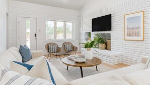 A spacious living room with a white brick fireplace, a flat-screen TV mounted above it, and two wicker chairs. - Twin Fin by Avantstay Walk to the Beach - Chic 30A Retreat (Panama City Beach)