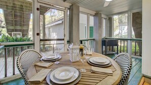 A covered outdoor dining area with a round table set for two, surrounded by wicker chairs, a white lantern, and a small table with a bottle and glasses. - Red Maple by Avantstay Walk to Folly Beach (Folly Beach)