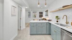 A modern kitchen with light blue cabinets, white countertops, and stainless steel appliances. There are pendant lights, a sink with a gold faucet, and a potted plant on the counter. - Red Maple by Avantstay Walk to Folly Beach (Folly Beach)
