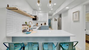 A modern kitchen with white cabinetry, a central island with bar stools, pendant lighting, and a view into the adjacent dining area. - Red Maple by Avantstay Walk to Folly Beach (Folly Beach)