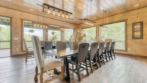 Ein Esszimmer aus Holz mit einem langen Tisch und Stühlen, großen Fenstern sowie einer Decke mit freiliegenden Balken und Pendelleuchten.