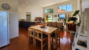 A spacious living area with a wooden dining table and chairs, a kitchen with a built-in oven, and a large window offering a view of the outdoors. - Easy Days - Ideal 2 wing set up, quiet street, central, 5min walk to the beach (Hahei)
