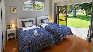 A modern bedroom with two beds, a nightstand, and a window with a view of a garden. - Easy Days - Ideal 2 wing set up, quiet street, central, 5min walk to the beach (Hahei)