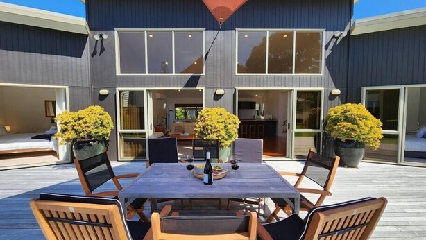 A modern house with a patio featuring a dining table and chairs, surrounded by large windows and potted plants. - Easy Days - Ideal 2 wing set up, quiet street, central, 5min walk to the beach (Hahei)