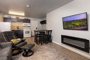 A modern living room with a fireplace, a flat-screen TV, a dining area with a table and chairs, and a kitchen in the background.