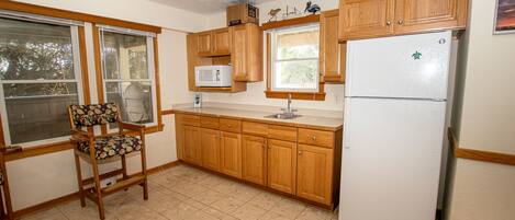 A kitchen with wooden cabinets, a white refrigerator, a microwave, and a window with a chair in front.