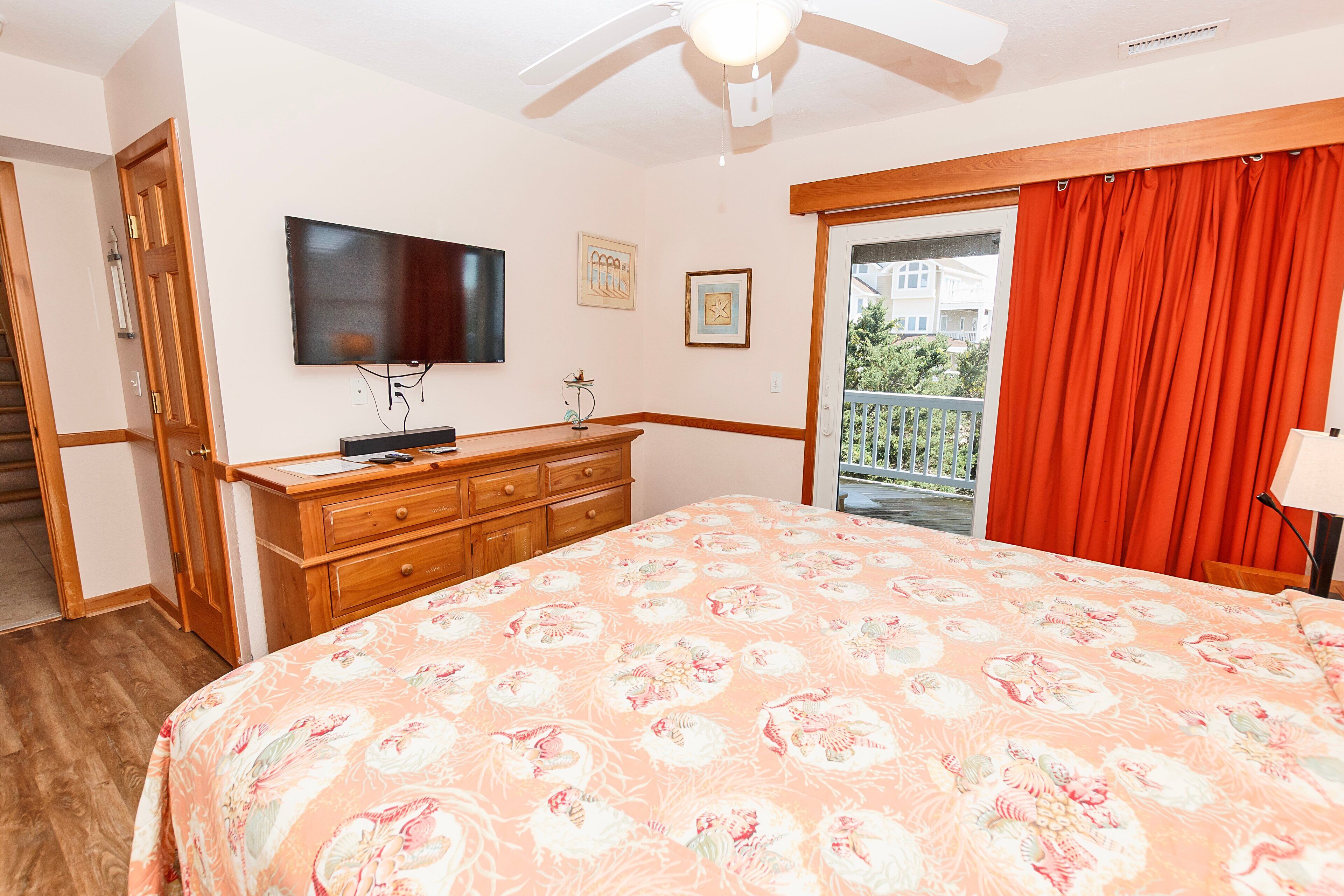 Un dormitorio con una cama grande, televisor montado en la pared, cómoda y balcón con cortinas rojas.