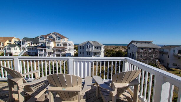A wooden deck with Adirondack chairs, overlooking a coastal residential area with multiple houses and a clear blue sky. - Semi-Oceanfront, Private Pool, Hot Tub, Ocean Views (Avon)
