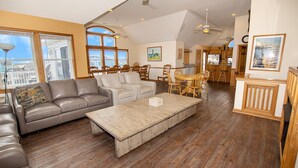 A spacious living area with a leather sofa, a wooden coffee table, and a dining table with chairs. There are large windows providing natural light and views of the outside. - Semi-Oceanfront, Private Pool, Hot Tub, Ocean Views (Avon)