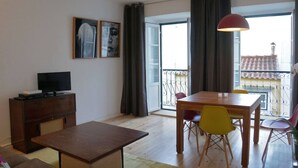 Een woonkamer met een houten tafel, stoelen, een televisie op een kast en een balkon met een hekwerk.