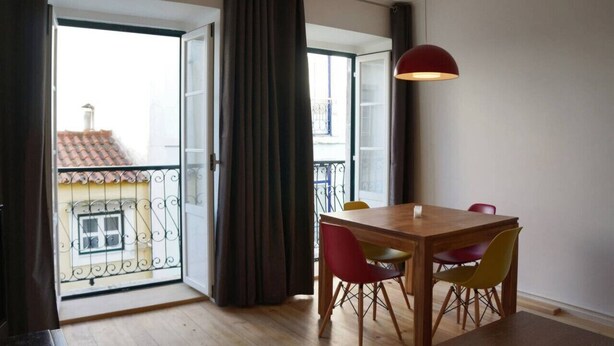 Een eethoek met een houten tafel en kleurrijke stoelen, een rode hanglamp, en uitzicht op een balkon met een hekwerk en een gebouw met een pannendak.