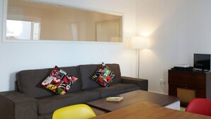 Een moderne woonkamer met een grijze bank, kleurrijke kussens, een houten salontafel, een rode fauteuil en een televisie op een houten kast.