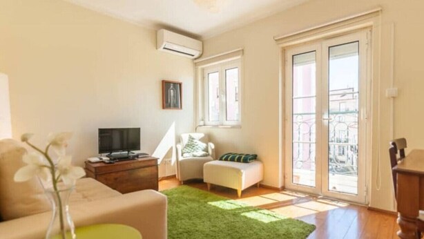 A living room with a sofa, a TV, a small table with a vase, a green rug, a chair, and a window with a view of buildings. - Cozy Two Bedroom Apt in Central Alfama | With A/c by Lisbeyond (Lisboa)