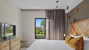 A modern hotel room with a large bed, a flat-screen TV, a window with a view of greenery, and a wooden headboard.