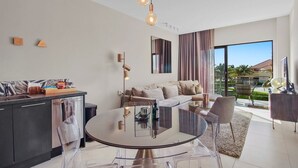 A modern living room with a glass dining table, a sofa, and a kitchen area with a sink and cabinets.