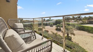 A balcony with wicker chairs and a view of a landscaped area with palm trees and a sandy beach.