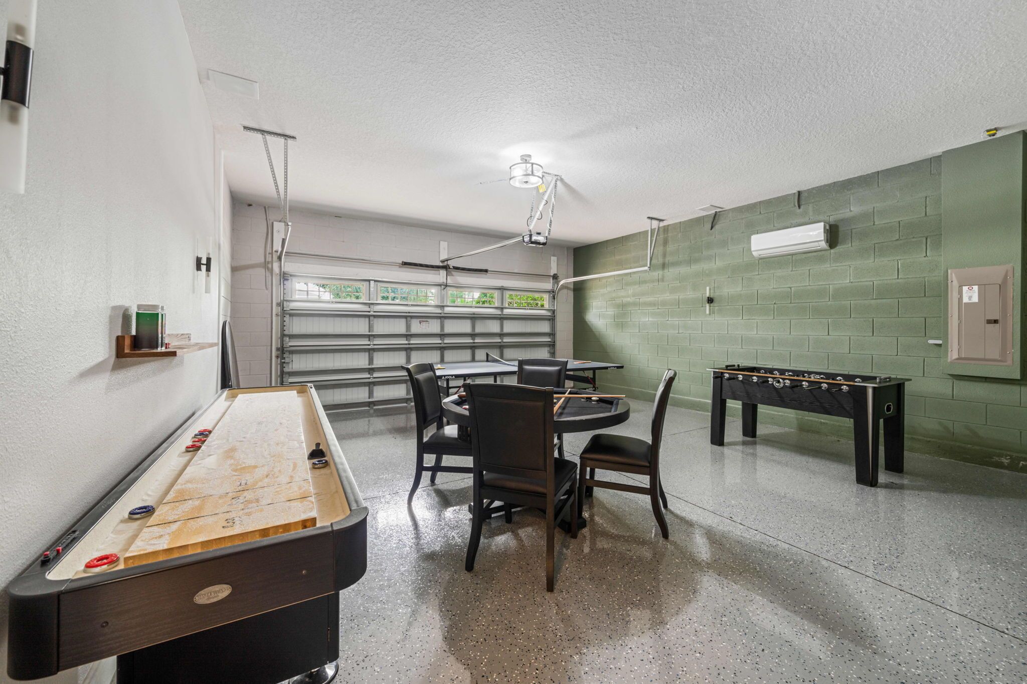 A pool table, chairs, a foosball table, and a table tennis table in a room with a green brick wall and a window.