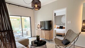 A modern living room with a sofa, a round coffee table, a wicker chair, a television on a wooden cabinet, and a large sliding glass door leading to an outdoor area.