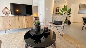 A modern living room with a flat-screen TV, a round coffee table with a candle, a wicker chair, and a wooden cabinet.