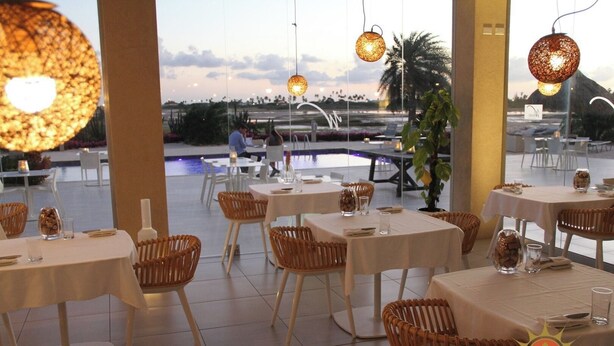 A restaurant with tables and wicker chairs, a pool in the background, and hanging lights.