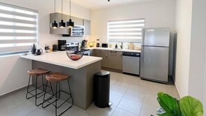 Et moderne køkken med en central ø, barstole, et rustfrit stålkøleskab og en sort skraldespand.
