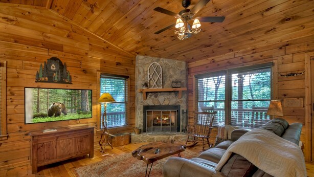 A cozy cabin interior with a stone fireplace, wooden walls, a leather sofa, and a television displaying a bear. - Cozy Retreat with Hot Tub, Firepit & Fireplace! - Bearfoot Ridge (Blue Ridge)