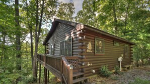 A log cabin with a wooden deck, surrounded by dense forest. - Cozy Retreat with Hot Tub, Firepit & Fireplace! - Bearfoot Ridge (Blue Ridge)