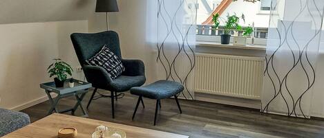 A modern living room with a dark green armchair, a small side table with a lamp, and a wooden coffee table. A large window with a view of a house and plants is visible.