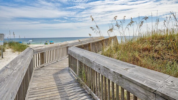 Ein Holzsteg führt zu einem Strand mit Sand und Meer im Hintergrund.