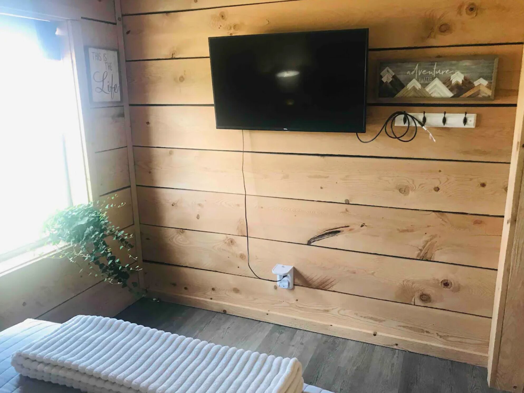 A wooden-paneled room with a flat-screen TV mounted on the wall, a small shelf with a 'Adventure Awaits' sign, and a power outlet on the lower wall.