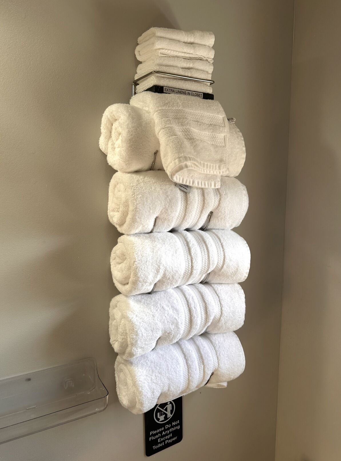 A stack of white towels on a wall-mounted rack with a sign below stating 'Please Do Not Flush Anything Except Toilet Paper'.