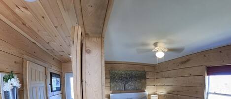 A wooden cabin bedroom with a bed, bedside lamps, a ceiling fan, and a window with a view.