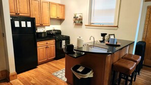 A kitchen with wooden cabinets, a black refrigerator, a stove, and a sink. There is a window with blinds and a small shelf with decorative items.