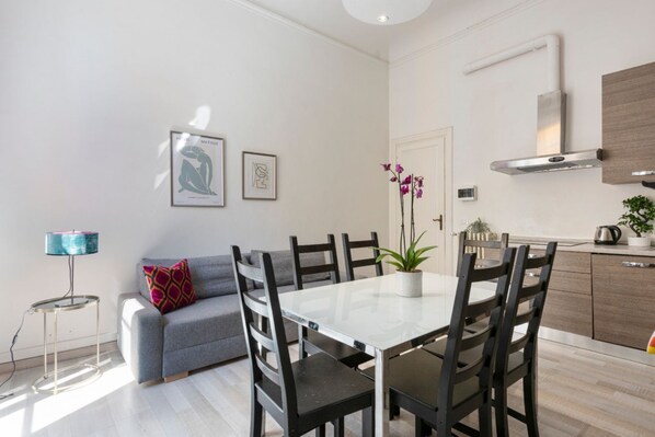 A modern living room with a dining table, chairs, a sofa, a small side table, a lamp, and a potted plant.