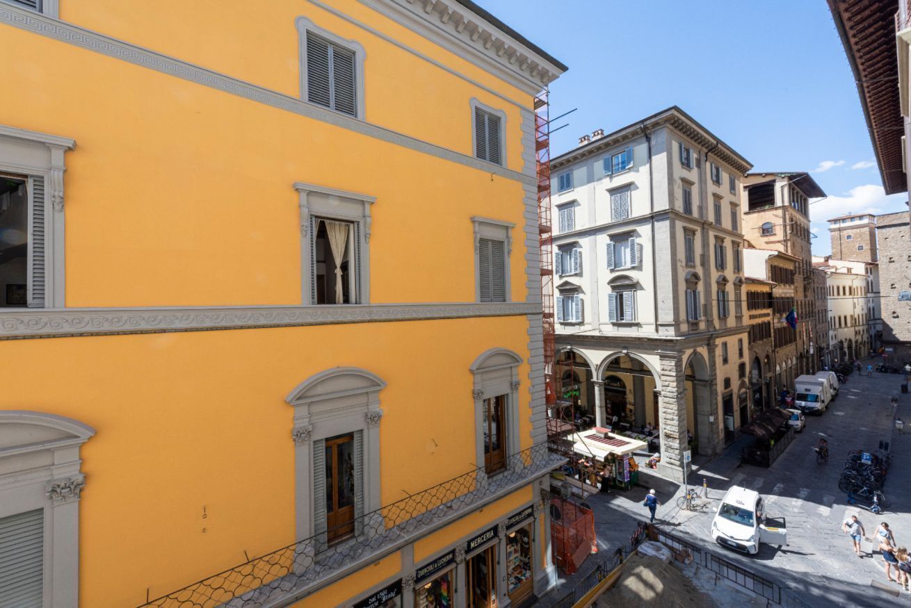 A yellow building with multiple windows and a balcony, adjacent to a street with pedestrians and parked vehicles.