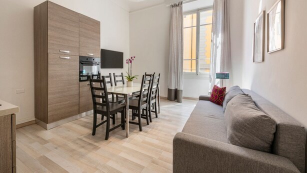 A modern living room with a sofa, a dining table with chairs, a TV mounted on the wall, and a window with curtains. - Apartments Florence - Pellicceria (sx) (Firenze)