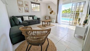 A modern living room with a sofa, a dining table, and a wicker chair. There are framed pictures on the wall, a window, and a view of a pool. - Villa Oneway with swimming pool - Rated 3 stars ⭐⭐⭐ (Sainte-Rose)