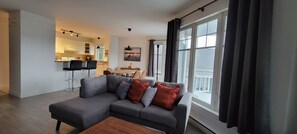 A modern living room with a grey sectional sofa, a wooden coffee table, and a dining area with a table and chairs. A kitchen is visible in the background.