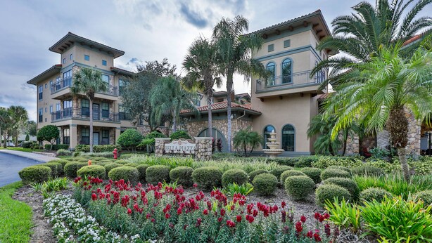 Un hotel de varios pisos con un jardín ajardinado que presenta flores rojas y blancas, palmeras y arbustos bien recortados.