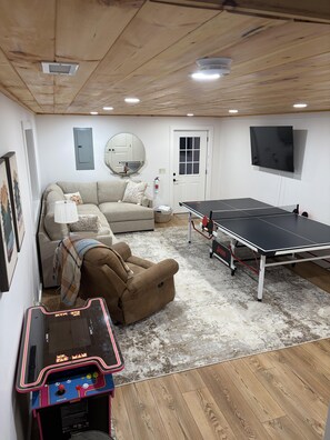 A room with a ping pong table, a sofa, a small table with a video game, and a mirror on the wall.